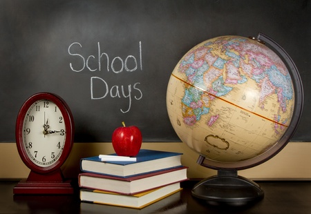 a chalkboard with the words school days written on it, a clock, books, apple, chalk and a globe sit on a teachers desk.の写真素材