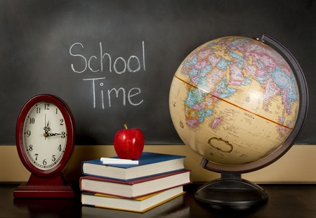 a chalkboard with the words school time written on it, a clock, books, apple, chalk and a globe sit on a teachers desk.の写真素材