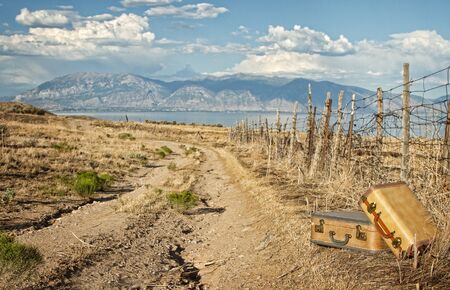 Vintage luggage sitting by a dirt road with mountains and lakeの写真素材
