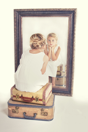 Child or young girl staring at herself in a mirror, sitting on vintage luggage, with a fish tail braid in her hair. Vintage or retro filter applied.