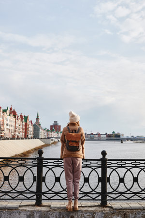Girl traveler with brown backpack enjoys the view of the city and river. The concept of travel, lifestyle, freedom. Embankment of Bruges. Yoshka-Ola, Mari El, Russia.の写真素材