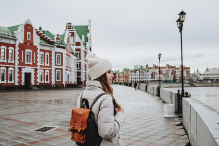 Traveler girl in a white jacket and a hat with a backpack enjoys a view of the Bruges embankment. Yoshkar-Ola, Mari El, Russia.の写真素材