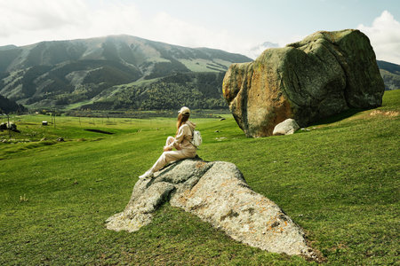 Young woman traveler sitting on rock in the middle of green meadow. Stone of desires in Semenovsky Gorge, Kyrgyzstan.の写真素材