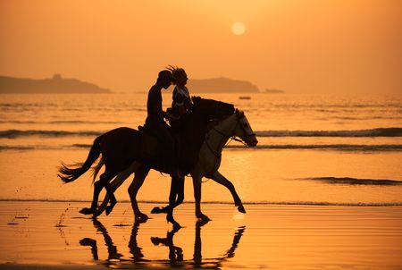 two people on horse back riding on wet sandy beach at sunsetの写真素材