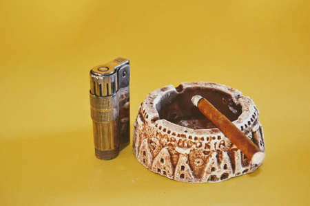 Retro and vintage ashtray and small type cuban cigar, metallic and retro lighter. Isolated yellow background. Smoldering cigarette or cuban cigar.の写真素材