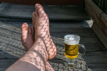 Feet of a resting man with a mug of light beer on a wooden floorの写真素材