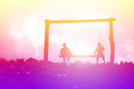 Silhouetted colorful scene of children sitting on swing bench of garden fieldの写真素材