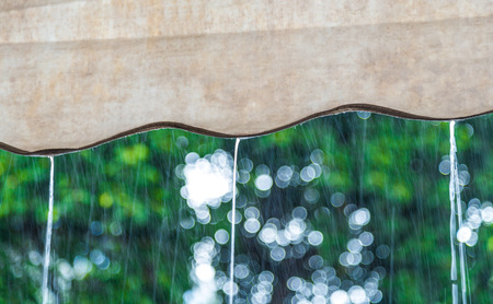 Close focus on rain flowing down from brown canvas roof. Outside is raining on blurry green tree background.の写真素材