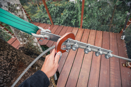 Hand holds safety ring lock on cable sling for walking on top tree wooden bridge.