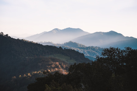 Mountain range in dark tone color covered by soft fog with white sky before sunset in winter season.の写真素材