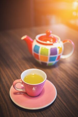 Close focus on pink ceramic teacup with green tea and colorful pot on table. Soft warm light from top corner.の写真素材