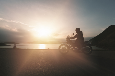 Silhouette scene of man riding motorbike on rough surface road near beach with golden sunshine from cloud of bright sky.の写真素材