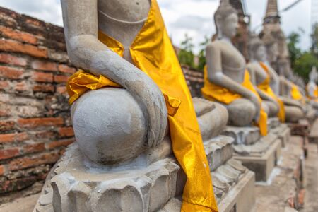 Close focus on hand of meditating monk statue in the row covered by yellow robe inside brick temple called Wat Yai Chai Mongkol in Ayutthaya of Thailandの写真素材