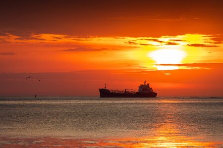 Silhouetted scene of ship and flying seagulls with cloudy orange sky at sunrise.の写真素材