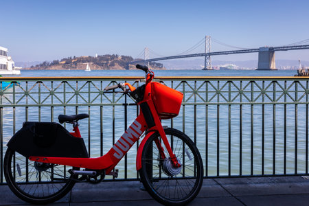 San Francisco, USA - September 27, 2019: Red rental bicycle painted 'Jump' on body parking on street near the fence at pier with background of San Francisco - Oakland Bay Bridge.のeditorial素材