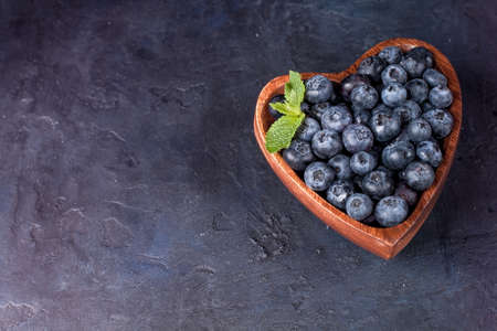 Fresh blueberries with a leaf of mint in the heart-shaped bowl on a dark blue background, flat layの写真素材