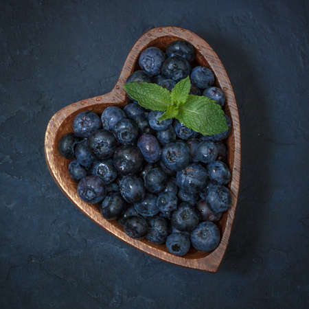 Fresh blueberries with a leaf of mint in the heart-shaped bowl on a dark blue backgroundの写真素材