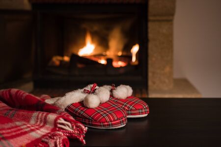 Wool warm blanket or plaid and red slippers with pompons on the background of burning fireplace....