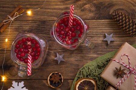 New Year Christmas composition with hot cranberry tea in glass mugs, net mesh bag with wrapped presents, cones, cinnamon, dried oranges and lights on wooden backgroundの写真素材