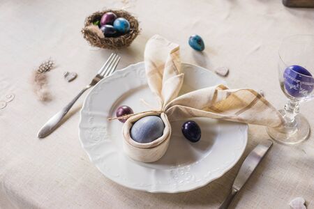 Easter festive spring table setting decoration, bunny ears shaped napkins, dyed hen's and quail eggs, feathers, wooden hearts, family dinner or breakfast conceptの写真素材