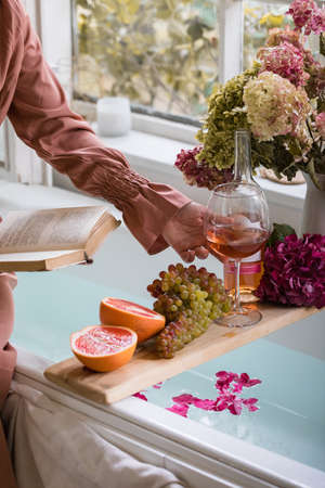 Bath tub with flower petals, grapefruit slices, bunch of grapes and woman's hand holding a glass of wine and a book. Organic spa relaxation preparationの写真素材