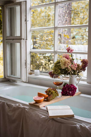 Bath tub with flower petals, grapefruit slices, bunch of grapes, a glass of wine, opened book and hydrangea bouquet. Organic spa relaxation preparationの写真素材