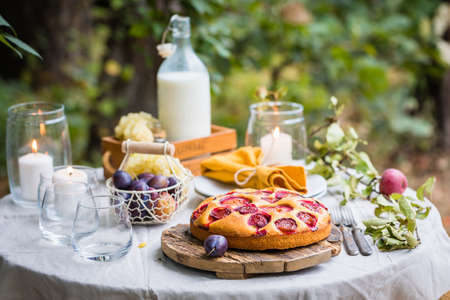 Traditional homemade plum cake or pie, summer table setting with milk, fruits, candlesの写真素材