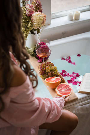 Bath tub with flower petals, grapefruit slices, bunch of grapes, woman sitting, a glass of wine and a book. Organic spa relaxation preparationの写真素材