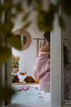Window outside view of Bath tub with flower petals, grapefruit slices, bunch of grapes, woman sitting, a glass of wine and a book. Organic spa relaxation preparationの写真素材