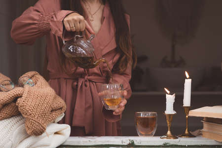 Warm and cozy autumn hygge concept with white and beige sweaters, woman pouring tea, burning candles and stack of old booksの写真素材