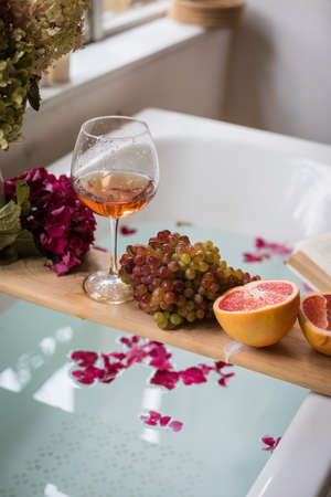 Bath tub with flower petals, grapefruit slices, bunch of grapes, a glass of wine, opened book and hydrangea bouquet.の写真素材
