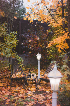 Fall themed calm garden holiday table arrangement with two glasses of red, books, maple leaves, lantern, wooden armchairs, tonedの写真素材
