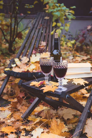 Fall themed calm garden holiday table arrangement with two glasses of red, books, maple leaves, lantern, wooden armchairs, tonedの写真素材