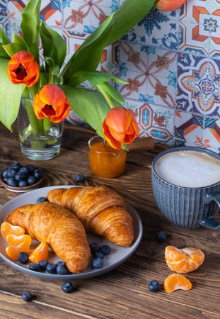 Breakfast with croissants, fresh blueberries, tangerines and coffee on the wooden kitchen table, orange tulips backgroundの写真素材