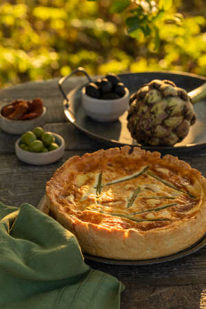 Golden organic healthy crispy pie quiche with artichokes, dried tomatoes and olives on the table, evening sunlight in the summer garden, soft selective focusの写真素材