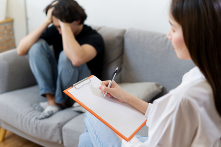Young male patient sitting on sofa closing his sad face is having consultant by professional psychologist female. Psychological consultant concept.の写真素材