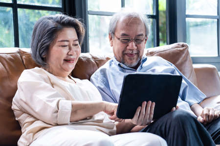 Asian loving senior elderly couple sit on sofa in living room use tablet read news together in...
