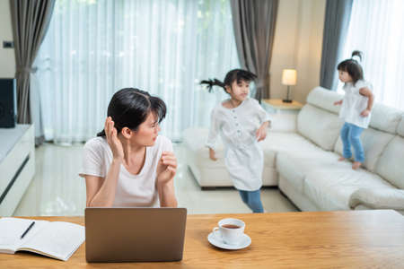 Asian businesswoman working at home due COVID-19 lockdown with children playing around. Freelance mother loss her concentration on job in computer feeling frustrated from young girl making noise.の写真素材