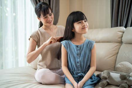 Asian loving mother spend leisure time with young little girl daughter in living room in house. Caring mom combing small child's hair with hairbrush with gentle. Family parenting relationship at homeの写真素材