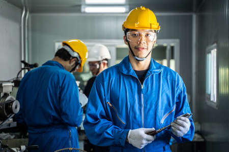 Portrait of Asian Mechanical worker smile, look at camera in factory. The Technicians male wearing protective glasses and yellow helmet for safety precaution, working and operating milling machine.の写真素材