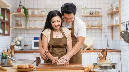 Asian new marriage couple stay home, spend time together in kitchen. Romance Young attractive husband and wife enjoy kneaded yeast dough with hand to bake bakery in house. Family activity relationshipの写真素材