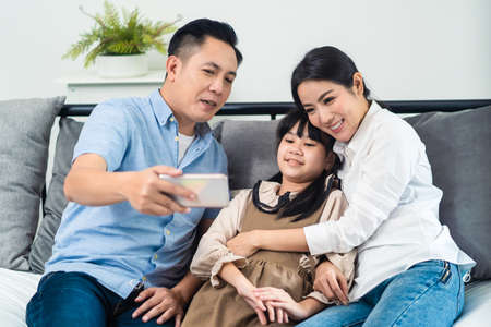 Asian Parents and little kid use telephone video call online to family. Loving Father Mother and young girl daughter sit on sofa in living room enjoy communicate with relative on smartphone in house.の写真素材