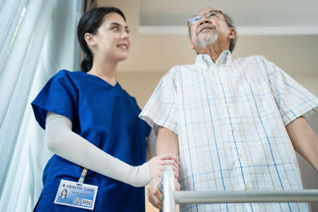 Asian Nurse support elder disable man walk with walker at nursing home. Caregiver therapist doctor help senior older handicapped patient use Zimmer frame in house. Medical therapy insurance concept.の写真素材