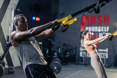 Group of multiethnic fitness trainer using ropes exercise in stadium. Active athlete fitness people man and woman lifting and push up on sling to maintain muscle and strengthen body at dark gym club.の写真素材