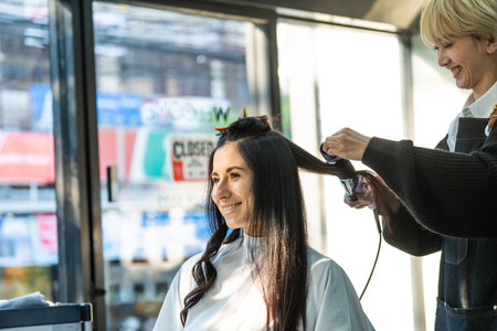 Asian stylist use curling iron make curly to customer's hair in salon. Caucasian beautiful girl with...