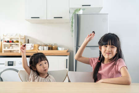 Portrait of Asian happy little cute kid holding chocolate in kitchen. Young lovely preschool child sibling sister sit on table enjoy eating sweet chocolate bar and smiling, looking at camera at home.の写真素材