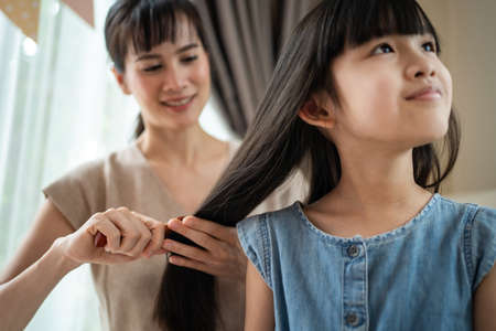 Asian mother combing little kid daughter's hair with hairbrush at home. Beautiful loving parent mom...