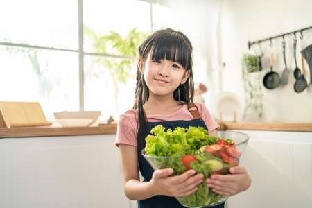 Portrait of Asian girl hold vegetable and look at camera in kitchen. Young attractive little kid child standing with happiness enjoy eating salad and healthy food for health care and wellness in houseの写真素材