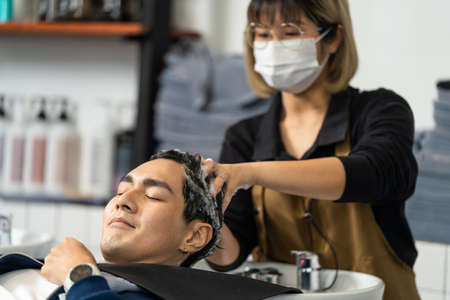 Asian attractive young male get hair washed by Hairdresser in salon. Expert hair stylist woman wear mask enjoy massage handsome man customer who's lying down on salon washing bed at beauty barber shopの写真素材