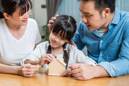 Asian Father and Mother teach young daughter to save money for future. Happy little girl enjoy put coins in piggy bank with parents and learn to manage financial plan. Loving Family couple concept.の写真素材
